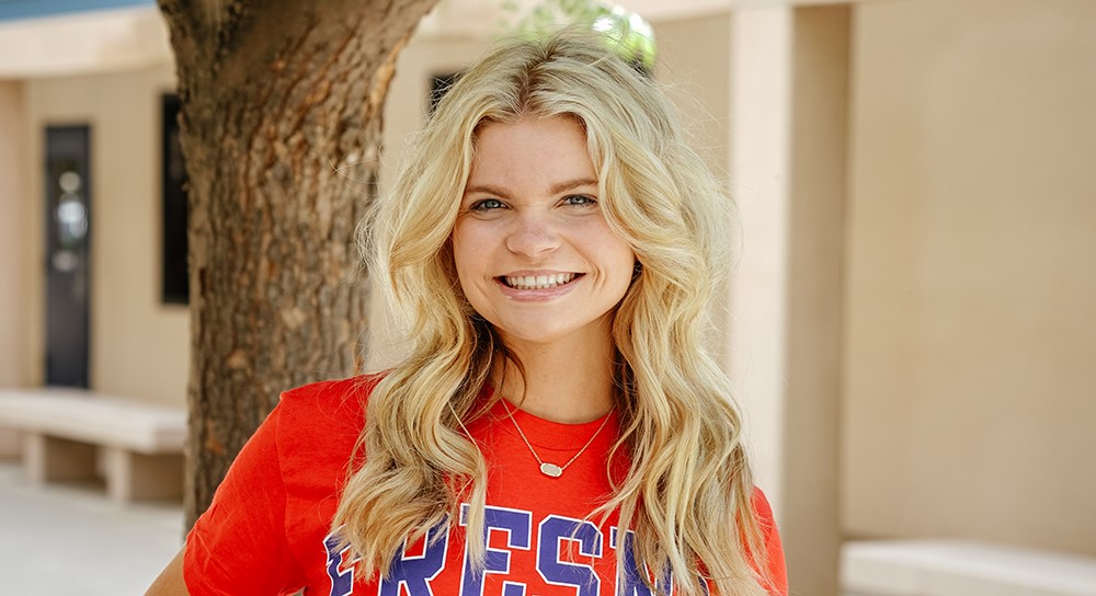 19-year old Trista Hulsey, with long blond hair and wearing a red Fresno State shirt, smiles while standing outside a building.