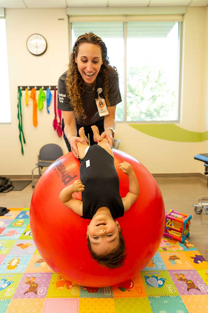 A healthcare worker holds a toddler boy by his legs as he lays back on a giant, inflatable ball. They are in a colorful, child-friendly rehabilitation room.