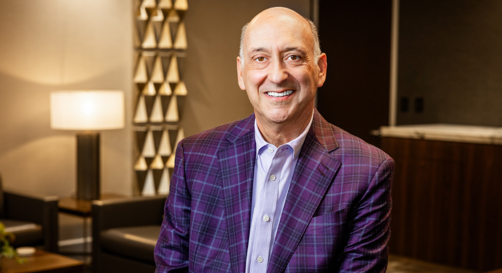 A man in a purple blazer sits in a comfortably lit office. He is smiling at the camera.