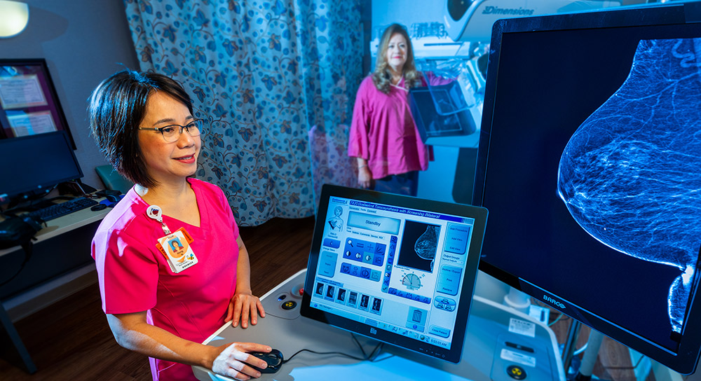 A healthcare worker in pink scrubs, standing at the left edge of the photo, smiles as she shows a screen with the diagnostic image of a breast. A woman in a pink gown stands in the background.