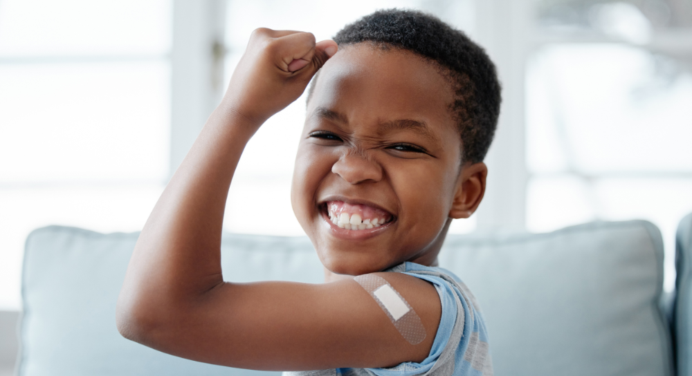 A young boy, about 7, flexes his arm and smiles at the camera. He has a band-aid on his bicep.