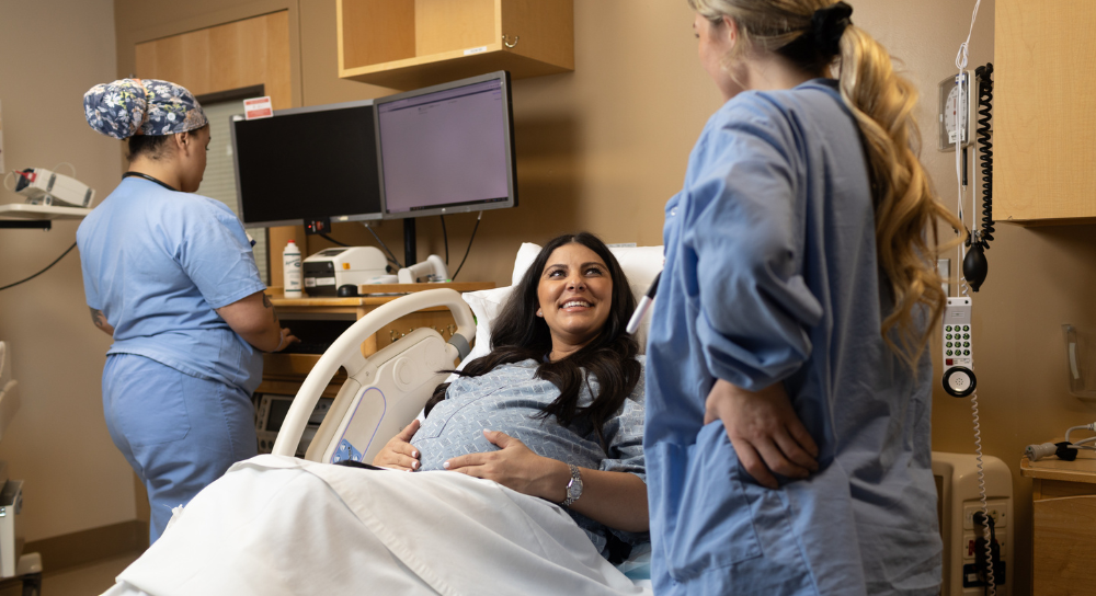 Three women in a maternity room. Two are healthcare workers. Their faces aren't visible. The third is a pregnant woman in bed who's smiling at one of the healthcare workers