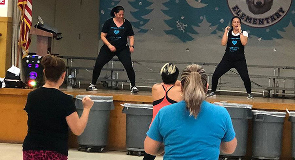 Two women stand on a school cafeteria stage and lead others in exercise.