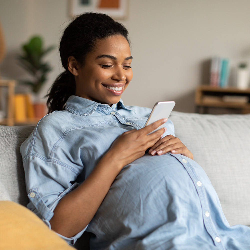 A young, Black mother looks smilingly at her phone as she rests her hand on her belly while sitting on a gray couch in a light, airy living room.