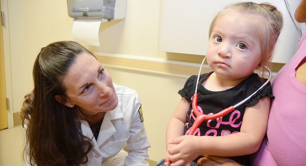 A UCSF doctor, white and female, bends down to smile at a toddler, female and Hispanic
