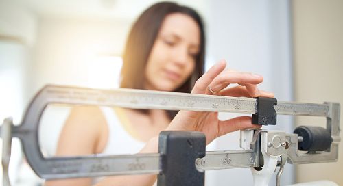 A young white woman adjusts the weights on an old-fashioned scale. The sun is shining brightly through the window behind her.