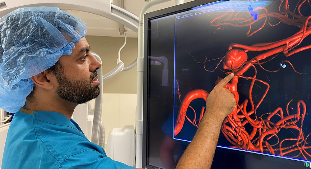 Dr Khan, in a scrub cap and blue scrubs, points to red lines on a black screen