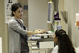 A nurse, female and Hispanic, types patient info into a computer. The patient's face is turned away, but she appears to be younger and Hispanic.