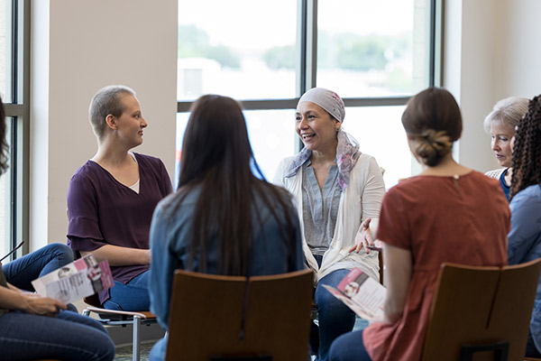 A group of women sit in a circle in chairs. Two have their backs to the camera. Two facing the camera have a shaved head and one wears a head scarf.