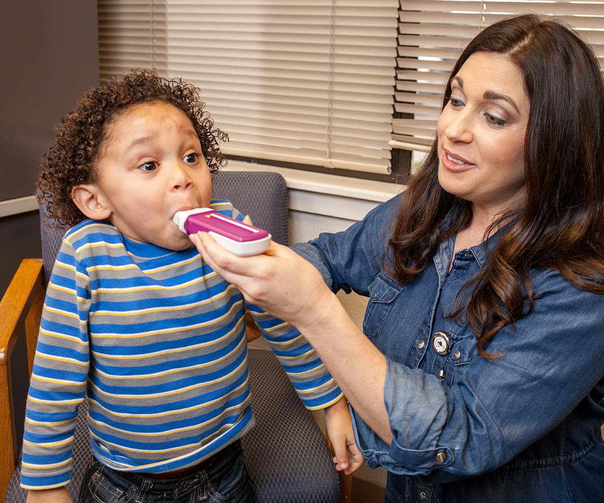 A toddler in a striped shirt with curly brown hair opens his eyes wide as a female healthcare worker helps him inhale an inhaler