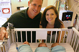 Fred and Kelsey Leyendekker pose with their triplets, who are swaddled and laying in a NICU bed
