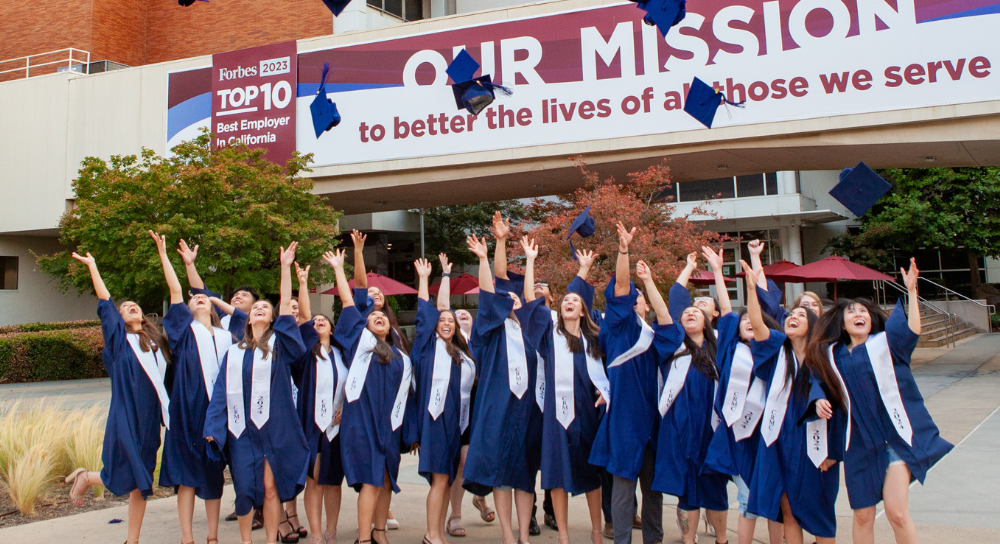 A group of 21 graduates in caps and gown toss their caps into the air.