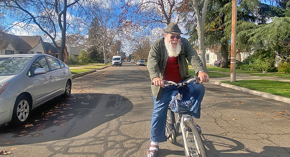 Paul Wilson, an older white man, rides his bike toward the camera with a blue sky behind him. 