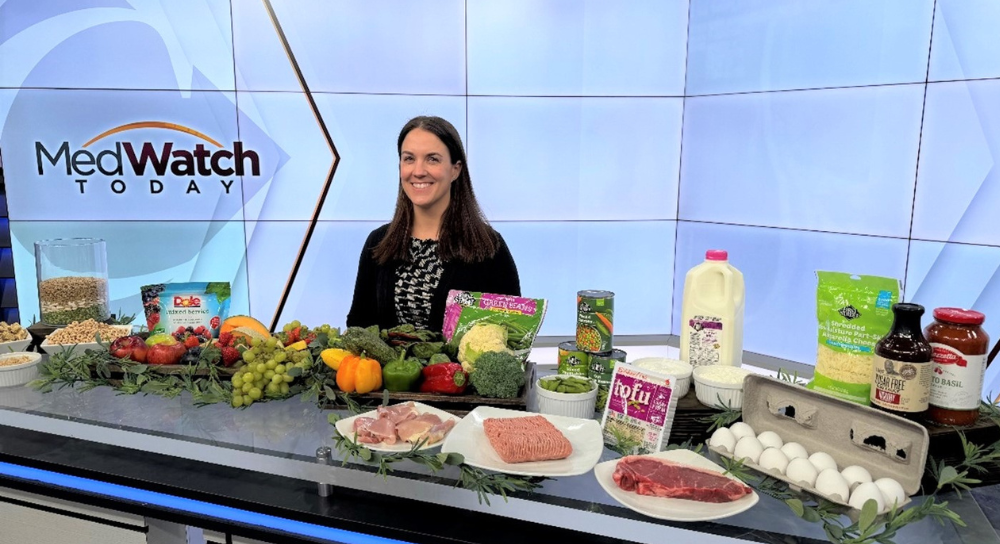 A smiling woman stands behind a table laden with nutritious foods.