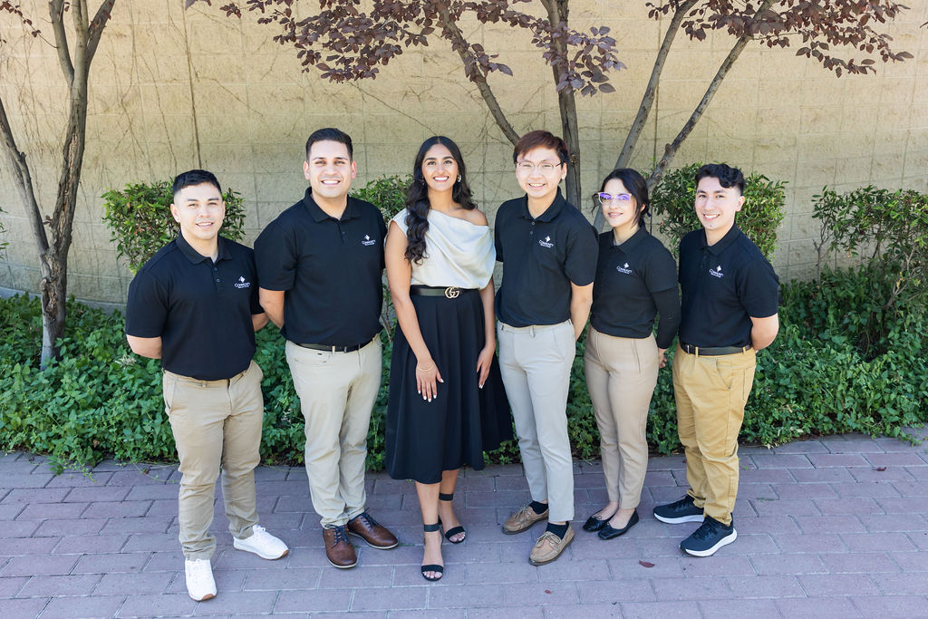 Six individuals stand on a sidewalk in front of a tree. Two are women, four men. They're posed and smiling for the camera.