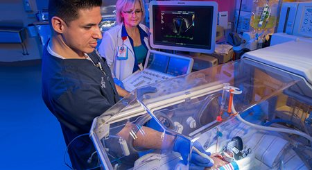 A male sonography student with a scanning device reaches into an incubator where a NICU patient sleeps. A blond woman, his teacher, watches from behind him.