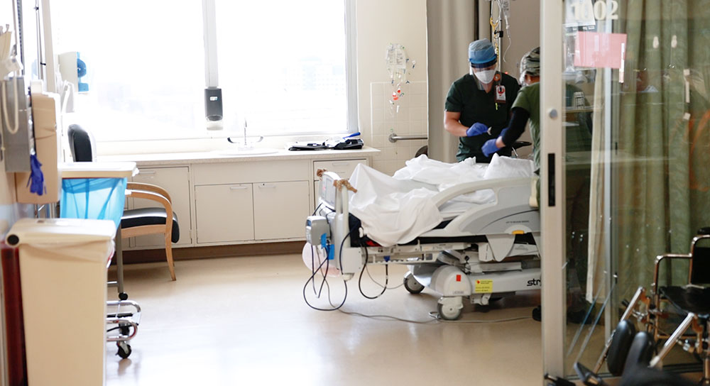 Wide shot of a hospital room with two masked healthcare providers attending to a patient whose face isn't visible