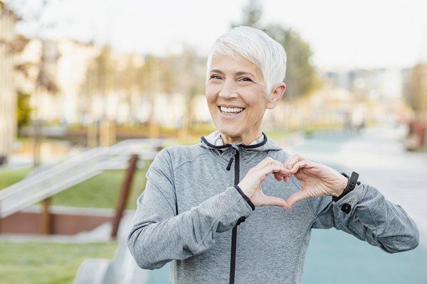 A mature white woman with white hair wear workout gear and forms a heart with her hands.