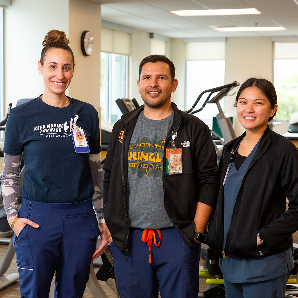 Three physical therapists stand in a gym: a white female, white male, and Asian female.