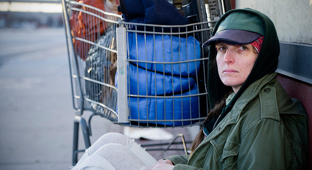 A white woman, houseless, sits on the ground and looks at the camera in distress.